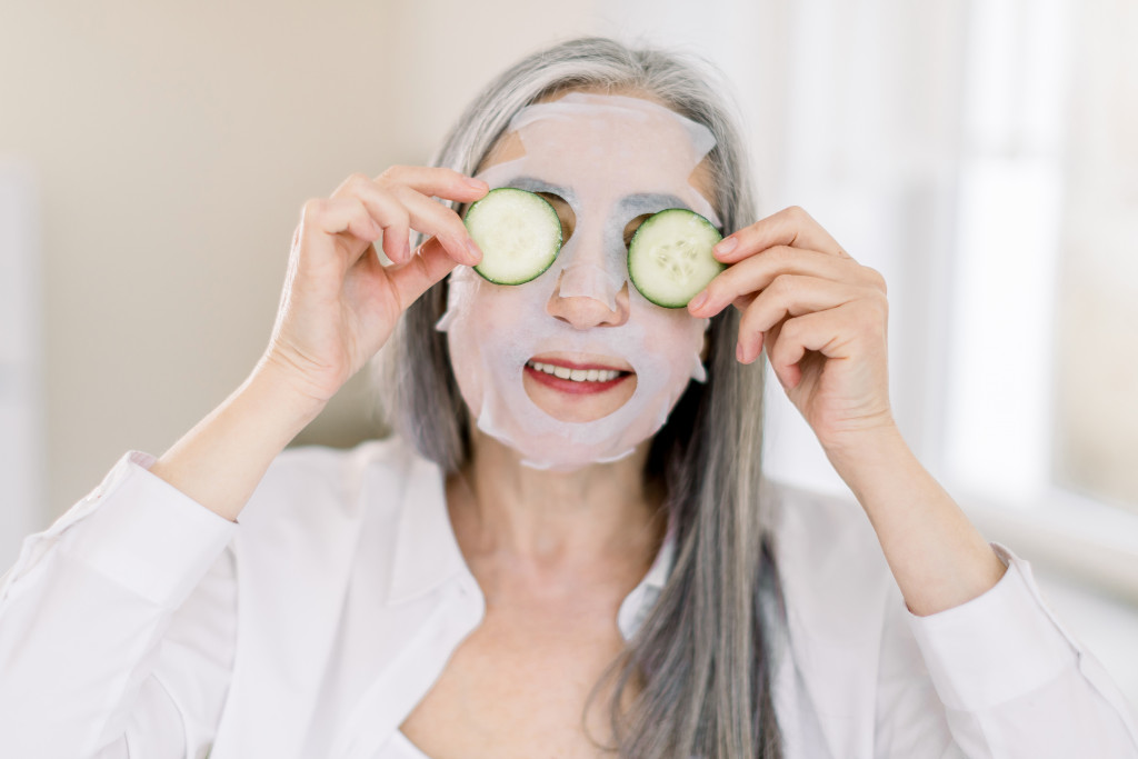 a senior woman with a cucumber while wearing a facial sheet mask